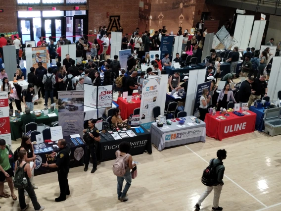 A college fair in a large hall filled with tables and banners and people visiting 